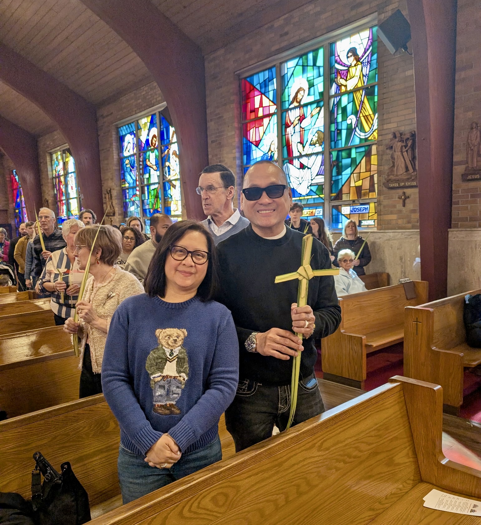 A couple standing in the pews holding a palm cross, in front of the church's stained glass windows
