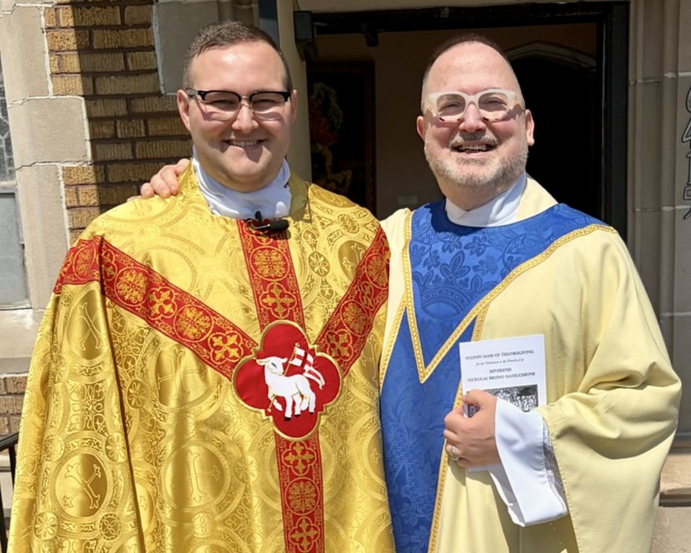 Two priests in gold and blue vestments stand arm in arm on the church steps