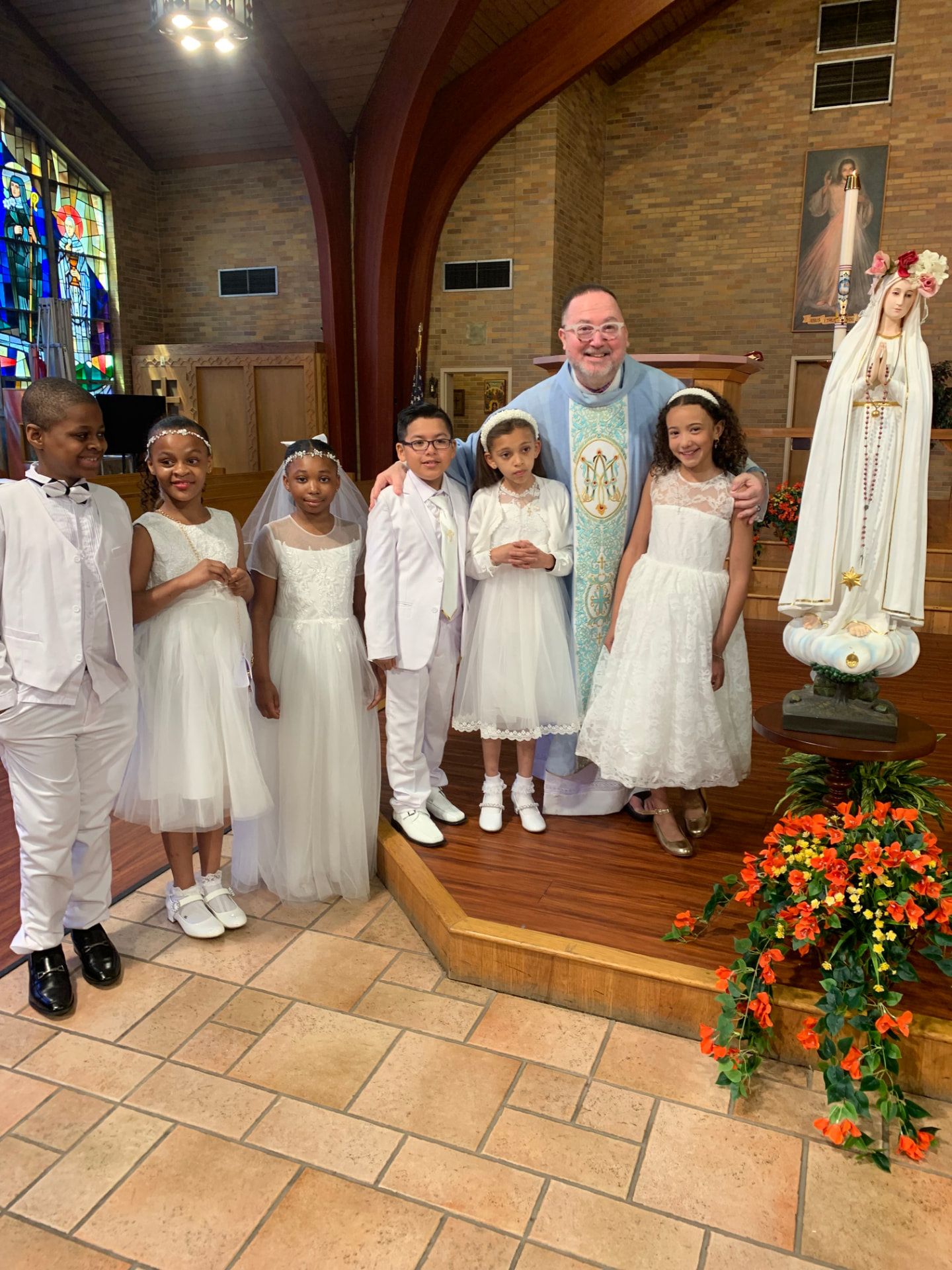 Fr. Jim with First Communion children in white dresses and suits beside the statue of Mary