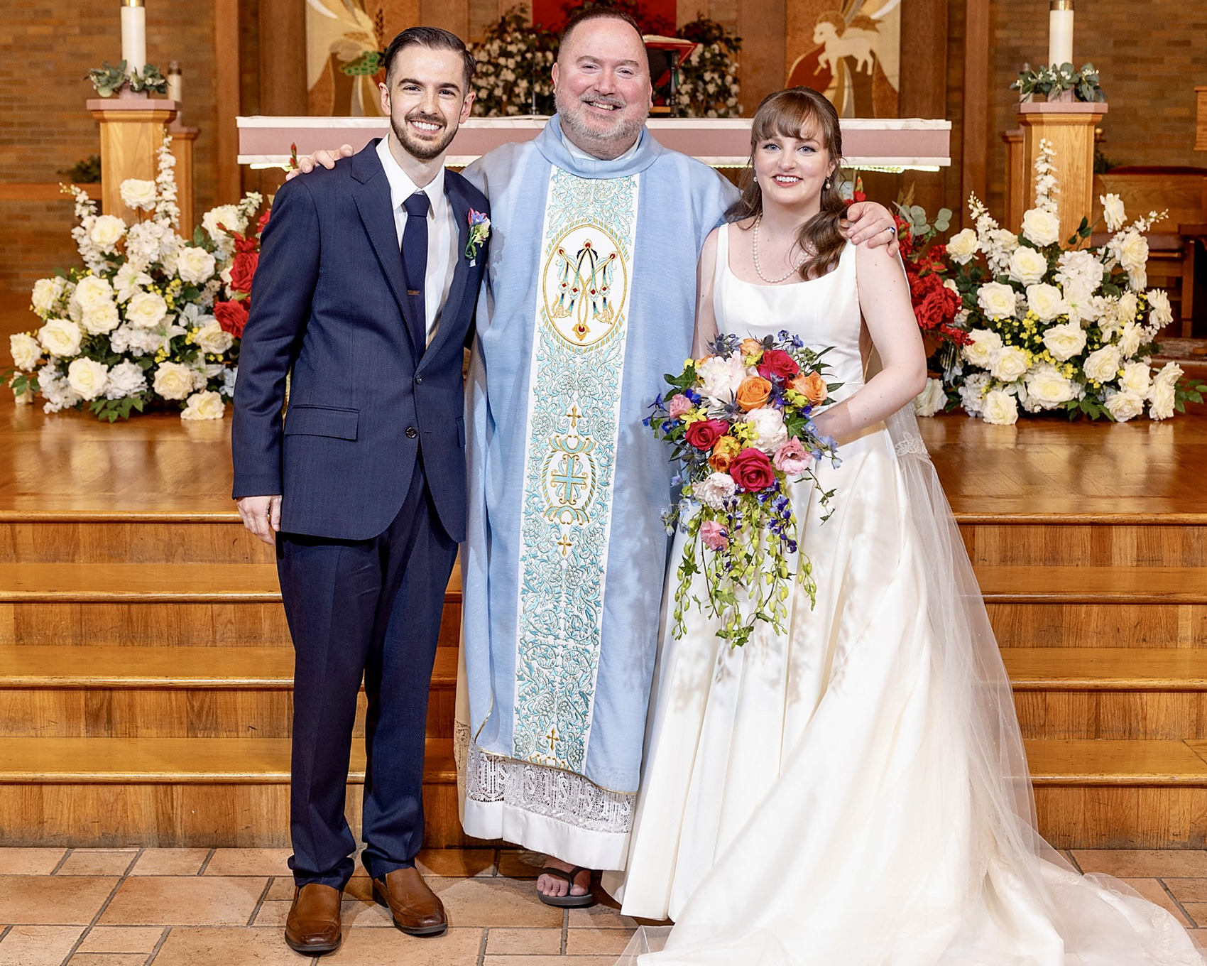 Bride, groom and priest pose at the altar steps with flowers and candles